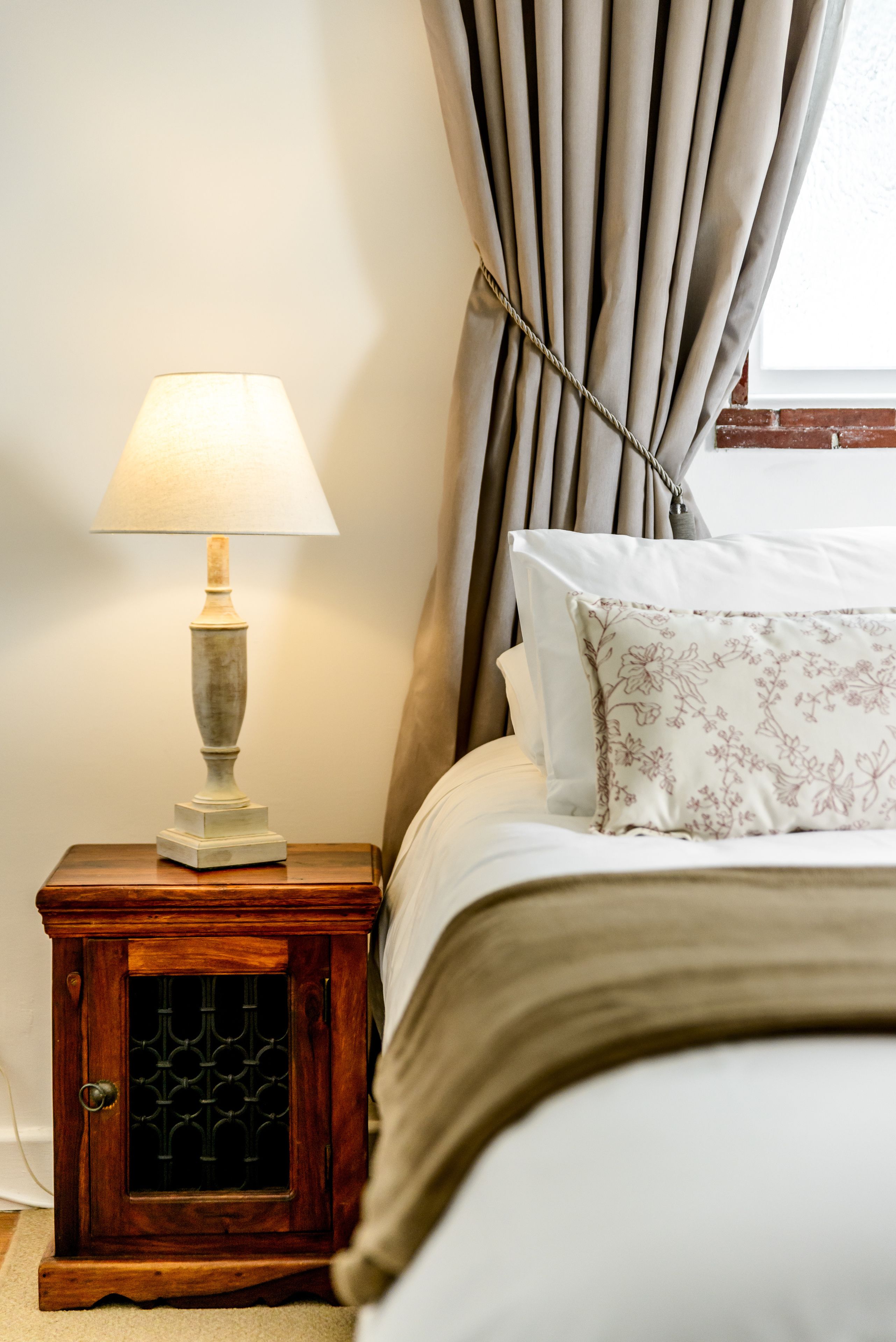 Un lit bien fait, agrémenté d’un coussin à motifs floraux, une lampe de chevet posée sur une table de nuit en bois et une fenêtre avec des rideaux.