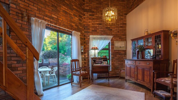 A room with a brick wall, wooden staircase, and a chandelier. There is a dining table with chairs, a cabinet, and a window with curtains. - Jenny's Guest House (Makhanda)