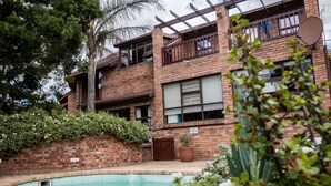 A two-story brick building with a swimming pool, surrounded by greenery and a palm tree. - Jenny's Guest House (Makhanda)