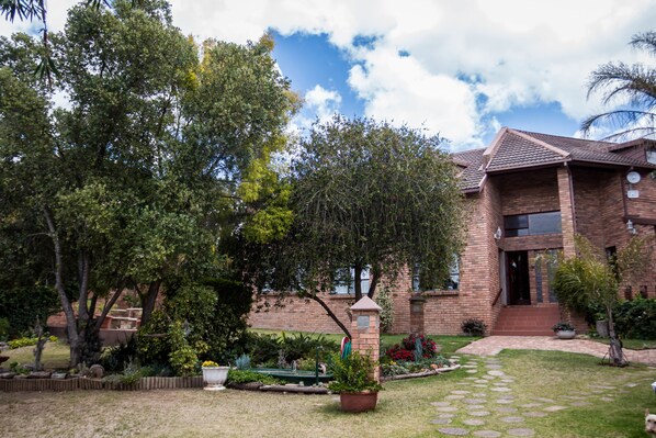 A brick house with a well-maintained garden, a stone pathway, and a large tree in the foreground. - Jenny's Guest House (Makhanda)