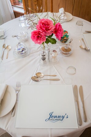 A round table set for a meal with a white tablecloth, clear glassware, and a centerpiece of pink flowers. - Jenny's Guest House (Makhanda)