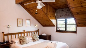A bedroom with a wooden ceiling, a bed with floral pillows, a nightstand, a bedside table, and a window with a view of greenery. - Jenny's Guest House (Makhanda)