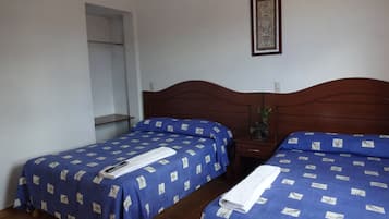 A room with two single beds, each with a blue bedspread and a white towel. There is a wooden headboard, a small table with a plant, and a framed picture on the wall.