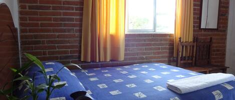A room with a brick wall, a bed with a blue bedspread, a window with curtains, a wooden chair, and a small table with a plant.