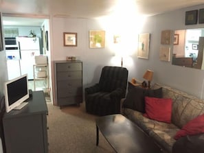 Una sala de estar con sofá, sillón y televisor sobre un mueble bajo. Se observan cuadros enmarcados en la pared y una lámpara sobre una mesita.