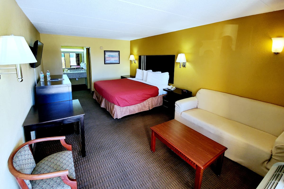 Ein Hotelzimmer mit einem Bett, einer Couch, einem Holztisch, einem Sessel, einem Schreibtisch mit einer Lampe, einem Fernseher und einem im Hintergrund sichtbaren Badezimmer.