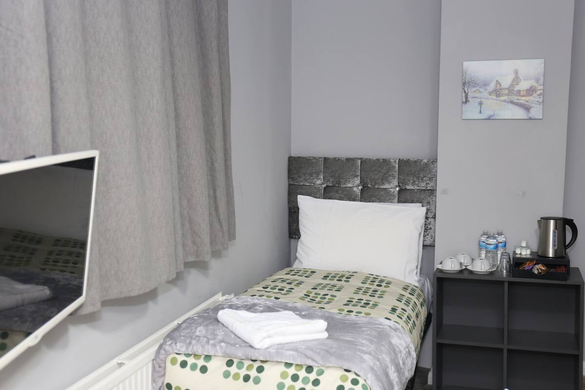 A neatly arranged bedroom with a bed, a bedside table with a kettle, and a painting on the wall.