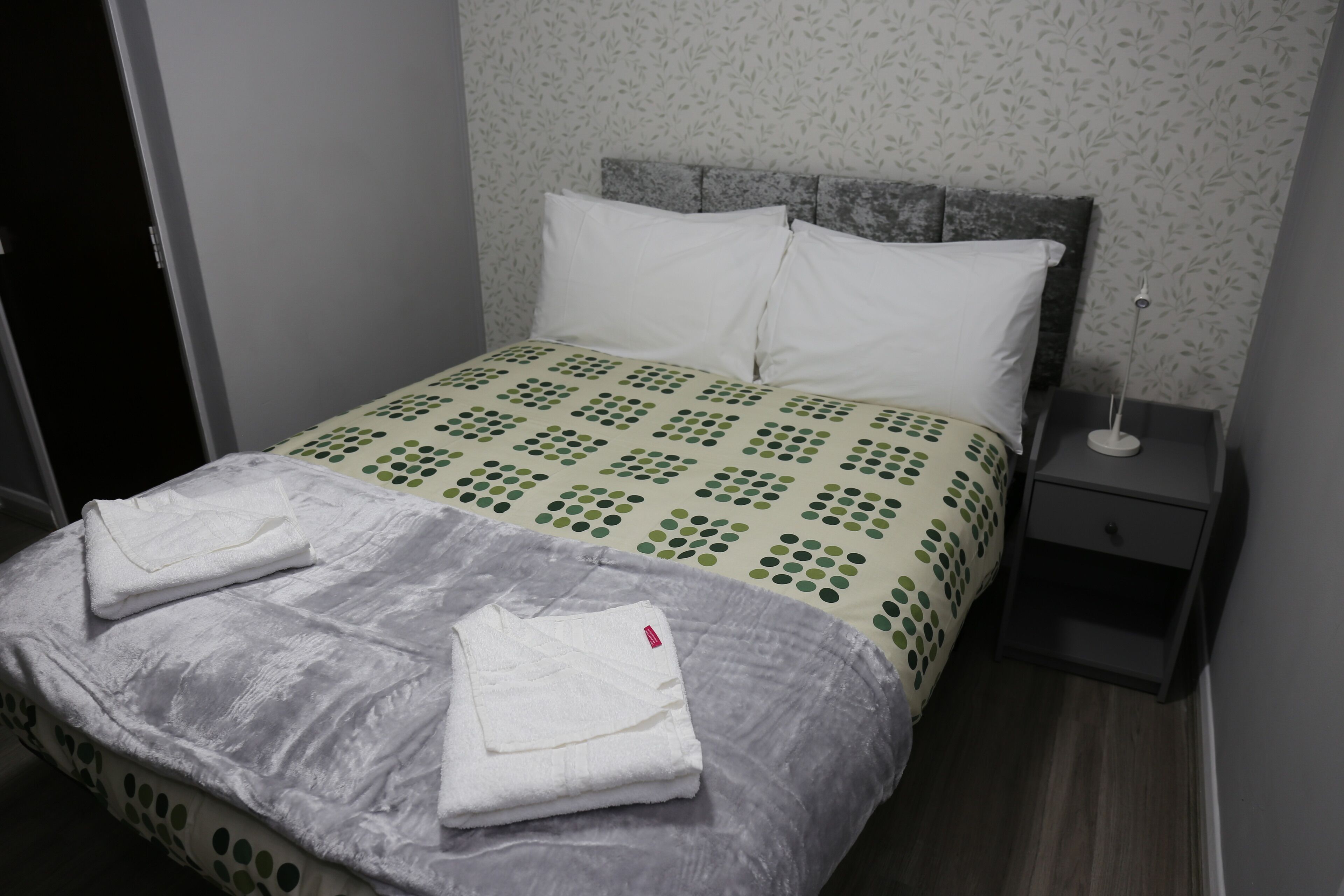 Una cama bien tendida con una colcha estampada, un cubrelecho gris y dos toallas blancas dobladas.