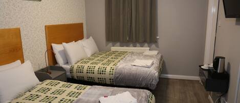 A hotel room with two beds, a wooden headboard, a grey patterned wall, a grey curtain, a television, and a small table with a kettle.