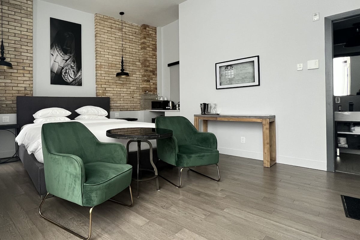 A modern hotel room with a brick accent wall, a bed with white linens, two green armchairs, a small round table, a wooden side table, and a black and white framed picture.