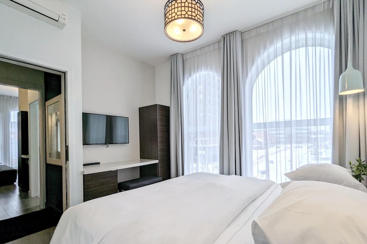 A modern hotel room with a large bed, a flat-screen TV mounted on the wall, a desk with a chair, and a large window with sheer curtains.