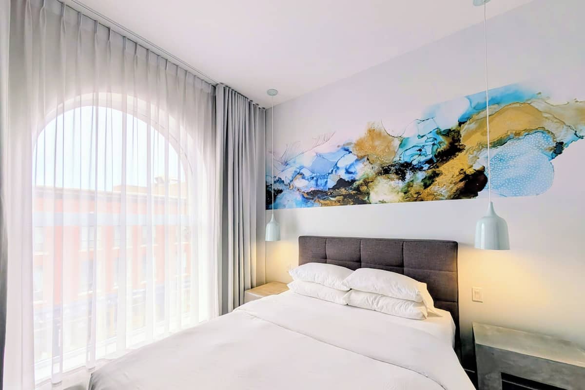 A hotel room with a large bed, a painting on the wall, and a view of buildings outside the window.