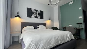 Una cama bien tendida con sábanas blancas, dos lámparas fijadas a la pared, un cuadro abstracto negro y un escritorio con una silla.