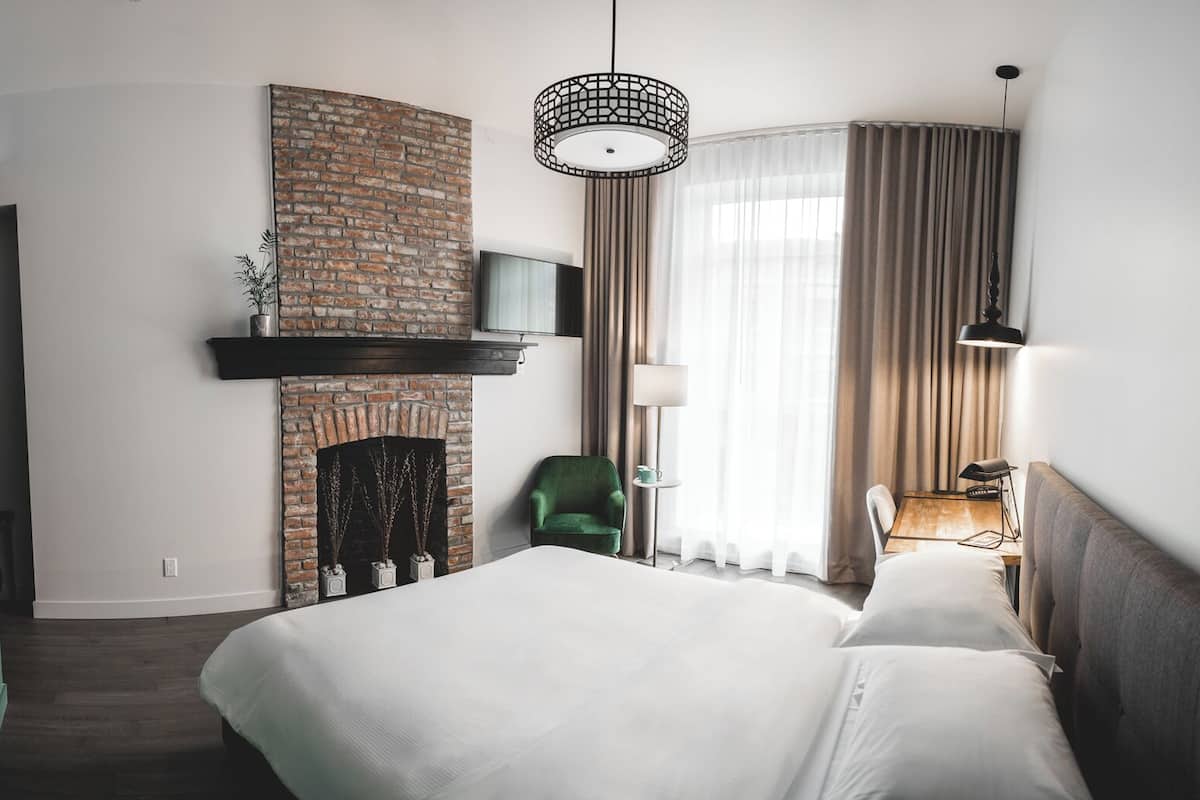 A hotel room with a brick fireplace, a bed, a green armchair, a desk with a lamp, and a large window with curtains.