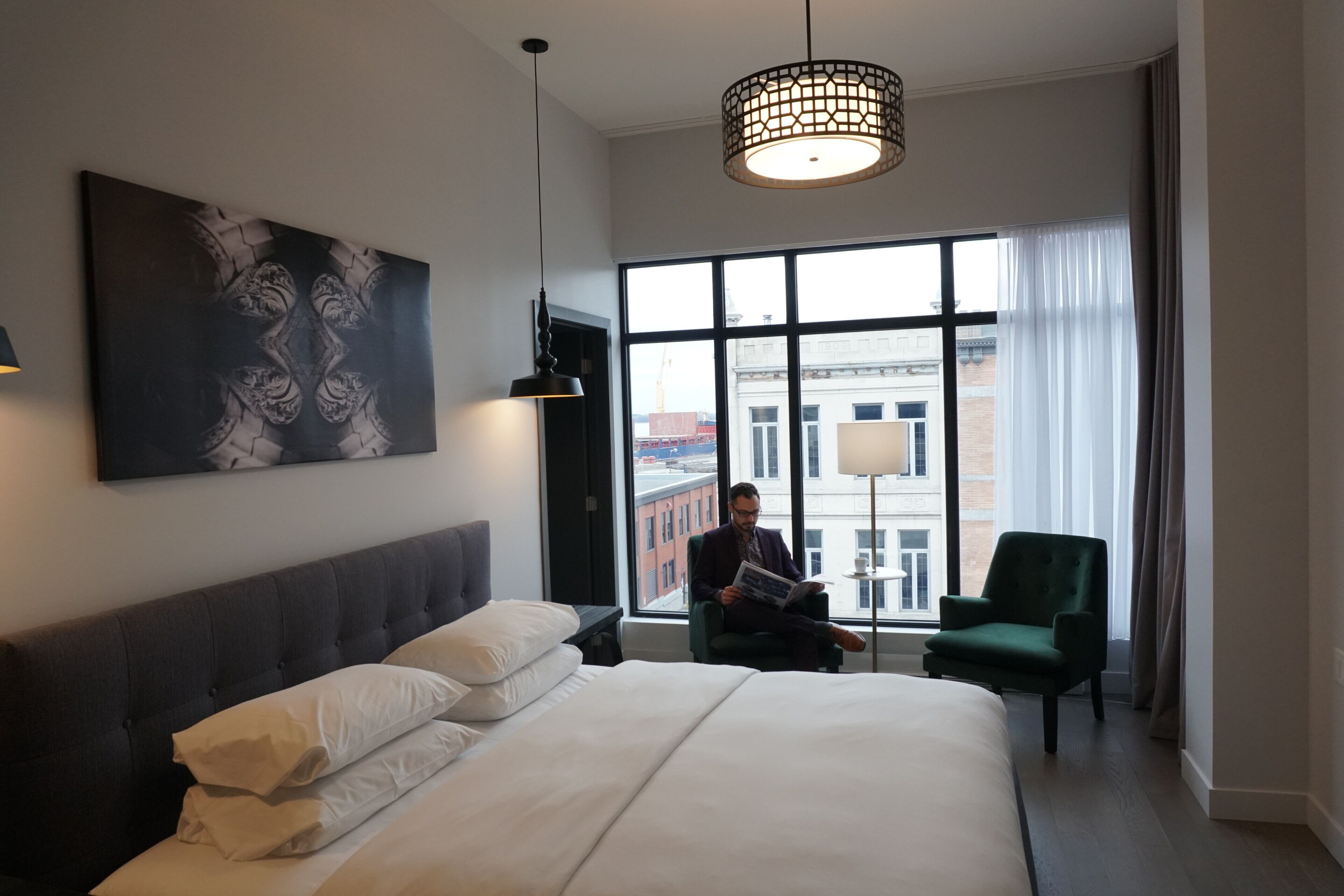 A hotel room with a large bed, a reading chair, a large window with a city view, and a modern pendant light.