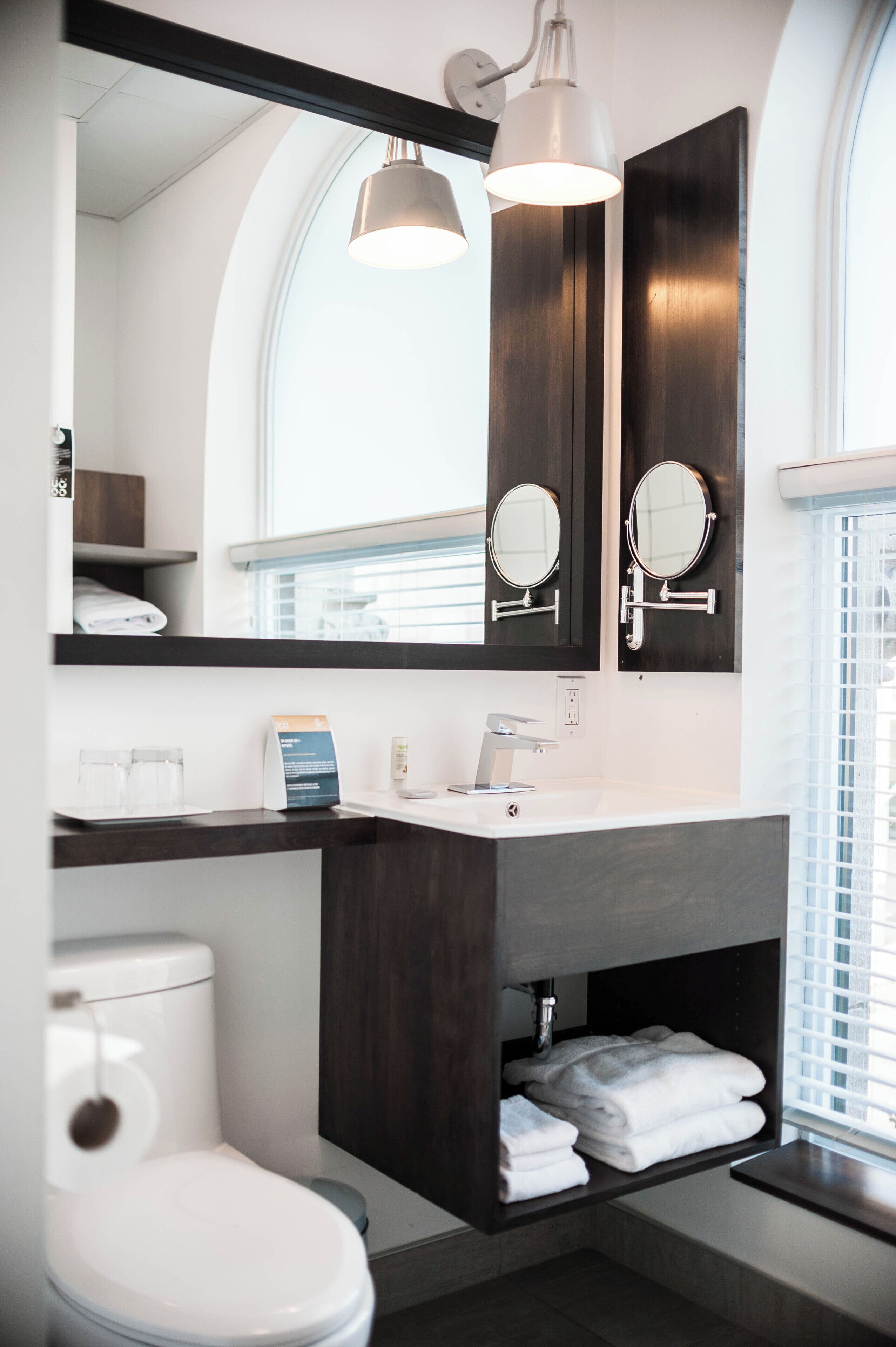 A modern bathroom with a toilet, sink, and two mirrors. There is a towel rack and a shelf with folded towels.
