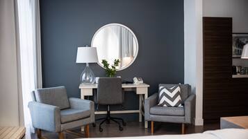 A modern hotel room with a grey armchair, a desk with a lamp, a round mirror, and a small plant.