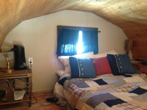 A cozy attic bedroom with a wooden ceiling, a bed with a quilt, a desk with a lamp, and a window with curtains. - Parlee Beach Luxury (Pointe-du-Chene)