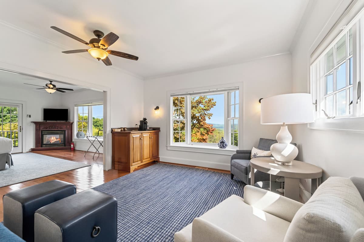 A spacious living room with a fireplace, large windows, and a ceiling fan.