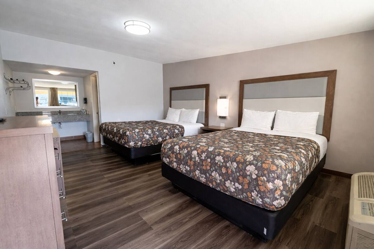 A hotel room with two beds, a wooden floor, a bathroom with a mirror, and a kitchenette.