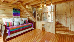 Interno di una baita in legno con una scala in legno, un divano con cuscini colorati e un arazzo intrecciato.