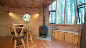 Interno di una baita in legno con stufa a legna, una finestra circolare a vetri colorati e vista sulla foresta attraverso ampie finestre.