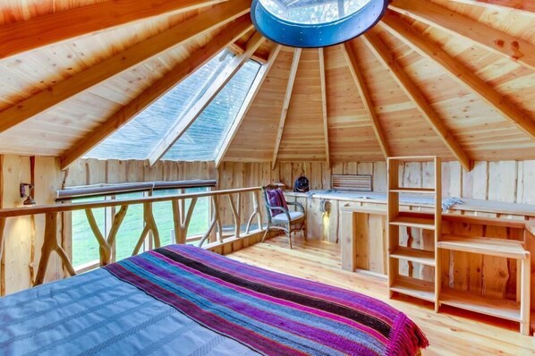 A wooden cabin with a sloped ceiling, a bed with a colorful blanket, a desk, and a chair.