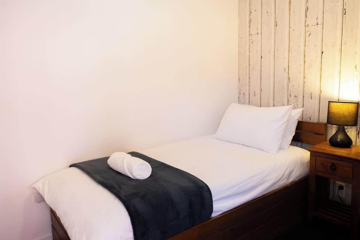 Ein Einzelbett mit weißer Bettwäsche und einer dunklen Tagesdecke, ein Nachttisch aus Holz mit einer Lampe sowie ein Holz-Kopfteil.