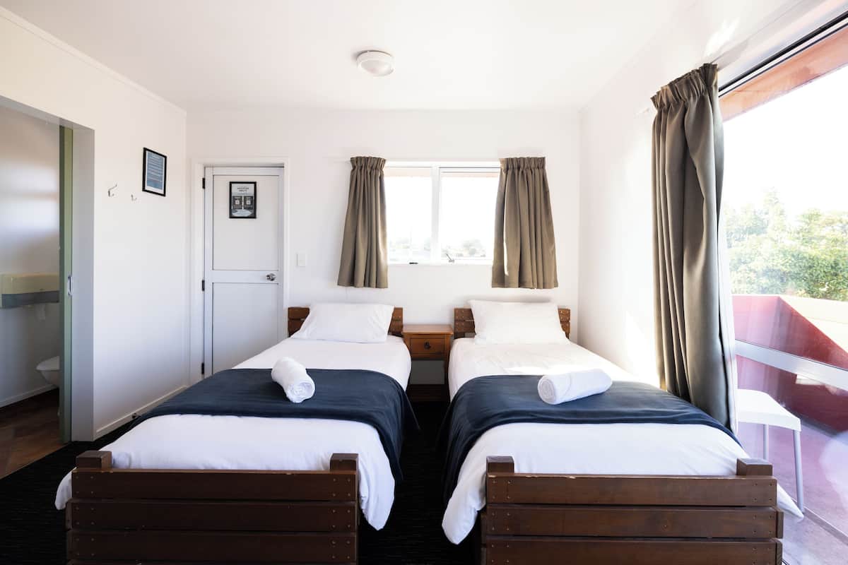 Ein Zimmer mit zwei hölzernen Betten, weißer Bettwäsche, einem Fenster mit Vorhängen, einer Tür und einem kleinen Nachttisch.