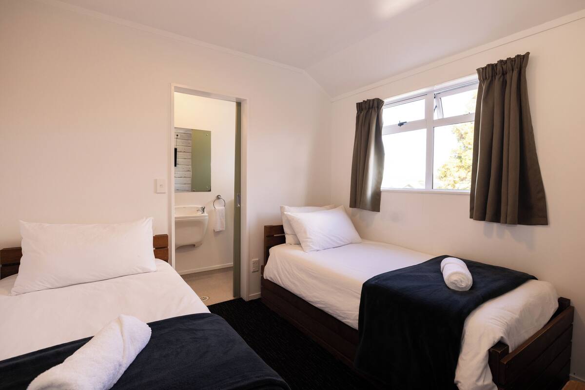 Ein Hotelzimmer mit zwei Einzelbetten, einem Fenster mit Vorhängen, einem sichtbaren Badezimmer und einer Tür zu einem weiteren Raum.