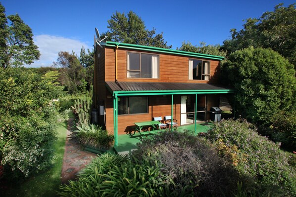 A modern two-story house with a green roof, large windows, and a patio area with outdoor furniture. - Capeview Cottage (Tirohanga)