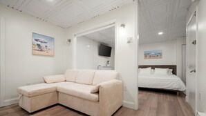 Una habitación con un sofá seccional color beige, una cama con ropa de cama blanca y un televisor montado en la pared.