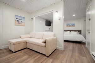 Una habitación con un sofá seccional color beige, una cama con ropa de cama blanca y un televisor montado en la pared.