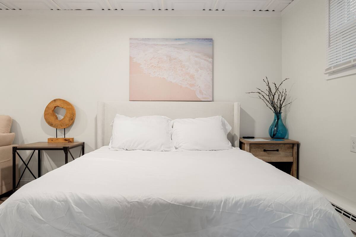 Une chambre à coucher comprenant un lit, une table de chevet, une commode, une œuvre d’art accrochée au mur et un vase avec des branches.