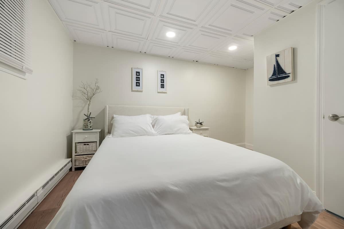 Une chambre avec un grand lit, des murs blancs, un plafond doté d’un éclairage encastré et une porte située à droite.