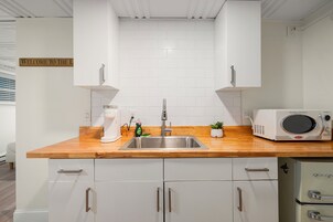 Una cocina moderna con armarios blancos, encimera de madera, pileta de acero inoxidable, microondas y una pequeña planta en maceta.