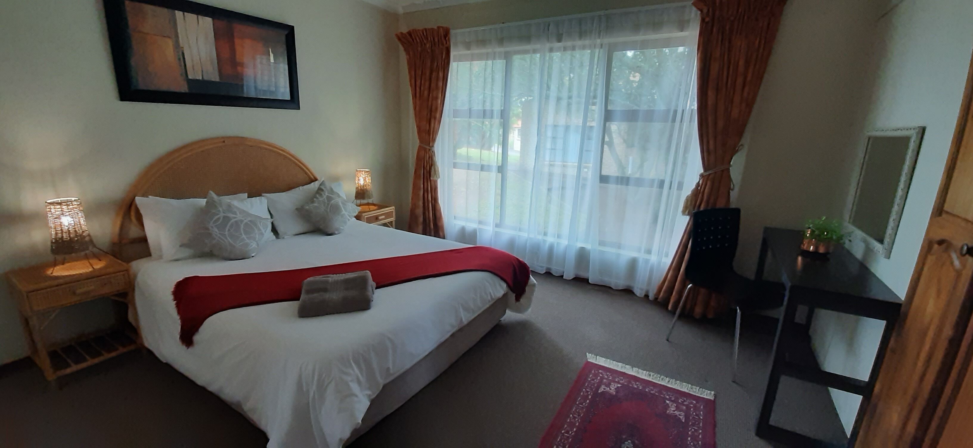 A neatly arranged hotel room with a large bed, a bedside table with a lamp, a TV, and a window with curtains.