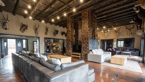 A spacious living room with a large gray sectional sofa, a white armchair, and a coffee table. The room features a rustic ceiling with exposed beams and a stone wall with mounted animal heads. - Humdani Game Lodge (Beestekraal)