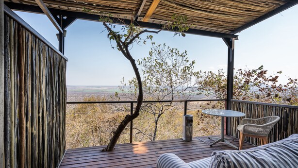 A wooden deck with a view of the landscape, a table, and a wicker chair. - Humdani Game Lodge (Beestekraal)