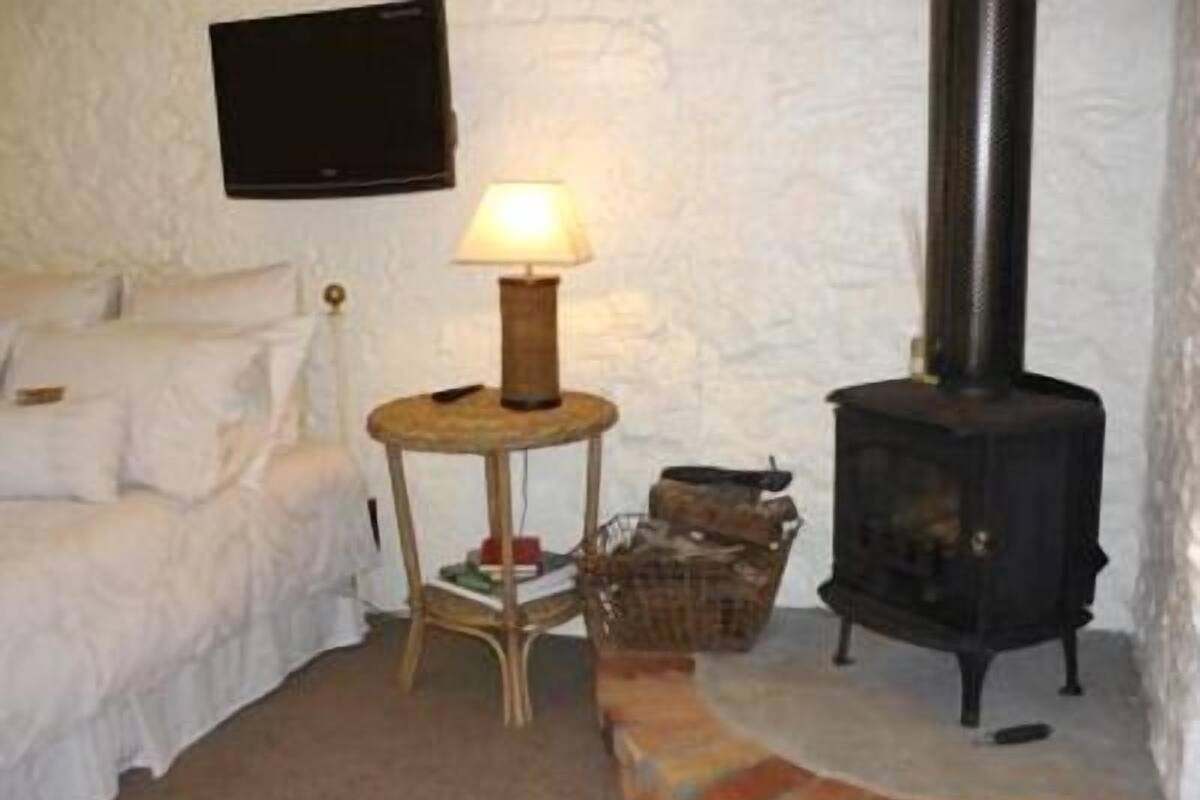 Une pièce chaleureuse avec un lit blanc, une petite table ronde surmontée d’une lampe et un poêle à bois noir.