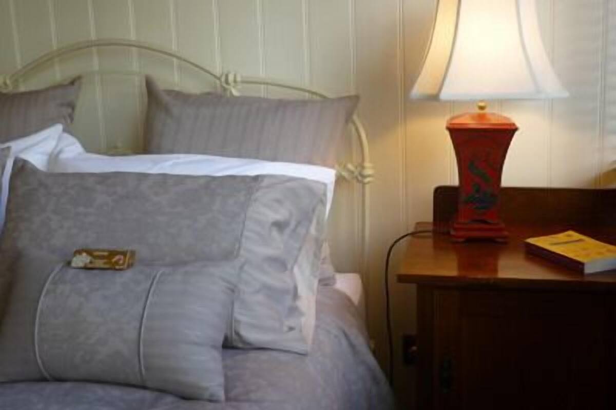 Un lit avec une couette et des oreillers gris, une lampe rouge posée sur une table de chevet en bois, et un livre sur la table de chevet.