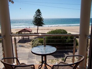 Un balcón con una mesa y sillas de vidrio, con vista a una playa que incluye un quiosco y un árbol solitario.