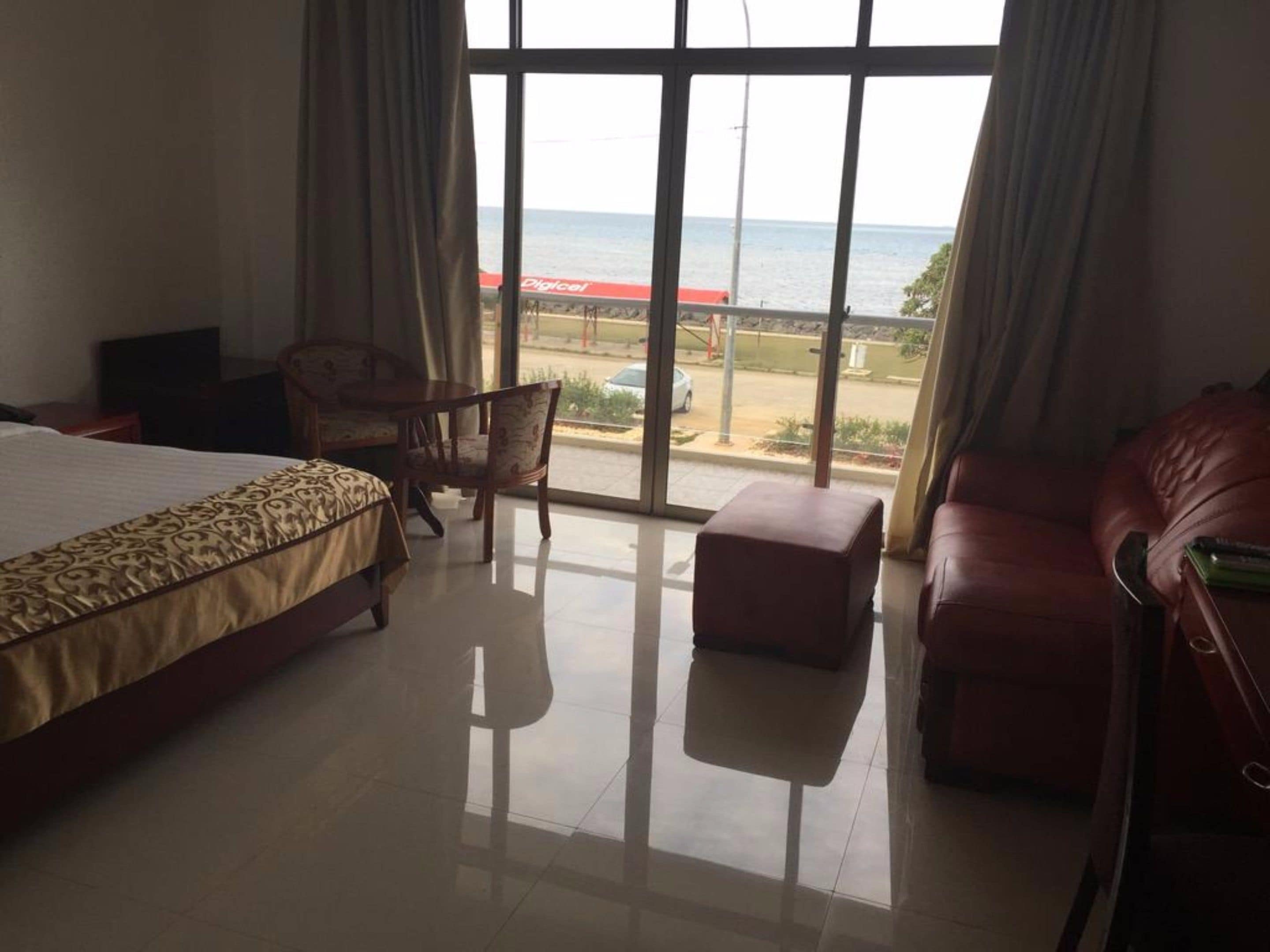 Habitación de hotel con una cama grande, una silla, un pequeño reposapiés y vista al mar a través de amplios ventanales.