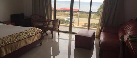 A hotel room with a large bed, a chair, a small ottoman, and a view of the sea through large windows.