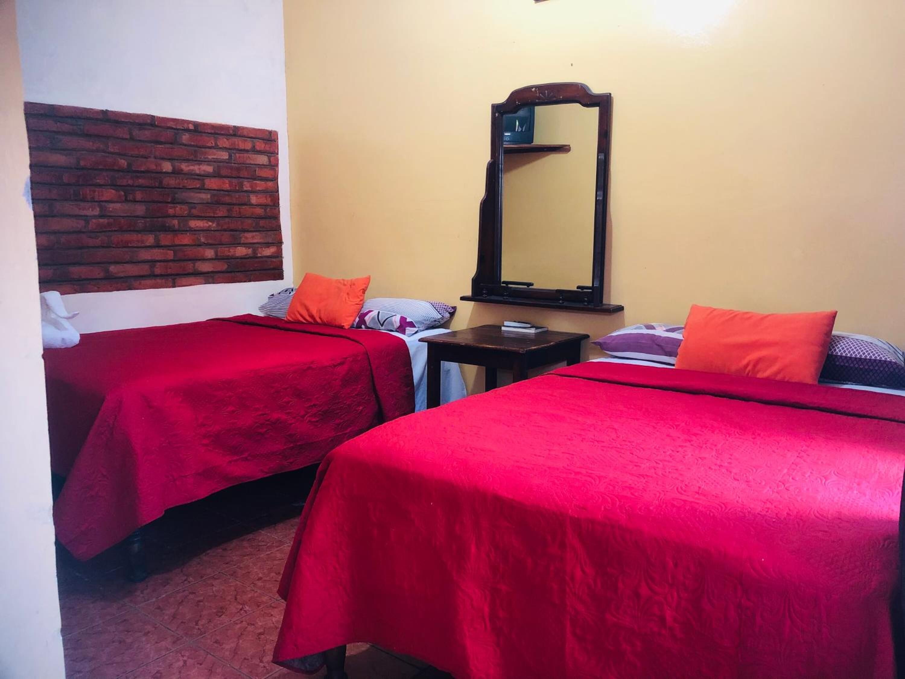 Una habitación con dos camas individuales, cada una con una colcha roja, una mesita con un libro y un espejo en la pared.