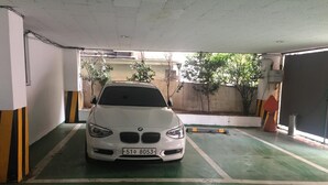 A white BMW parked in a parking garage with green and white painted lines, concrete pillars, and a view of trees and buildings outside. - Prince Motel (Gwangju)