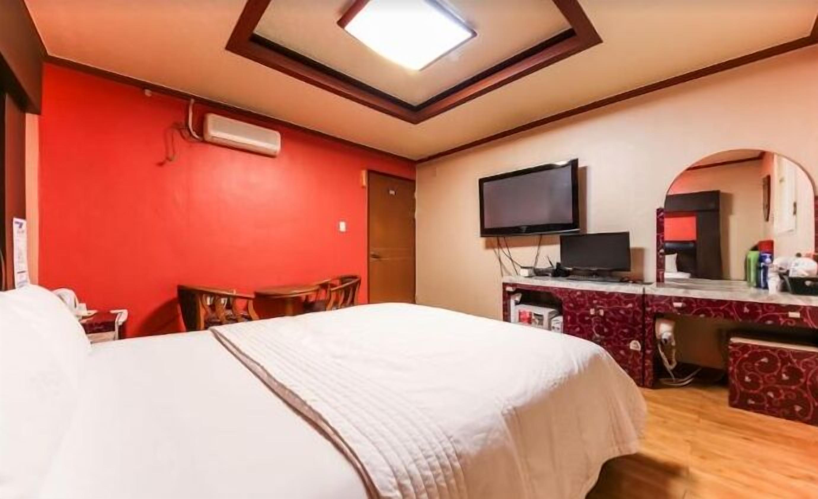 Habitación de hotel con una cama grande, un televisor, un escritorio con una computadora y una pared roja.