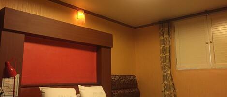 A hotel room with a bed, a headboard, a chair, a window with curtains, and a wall-mounted light fixture.