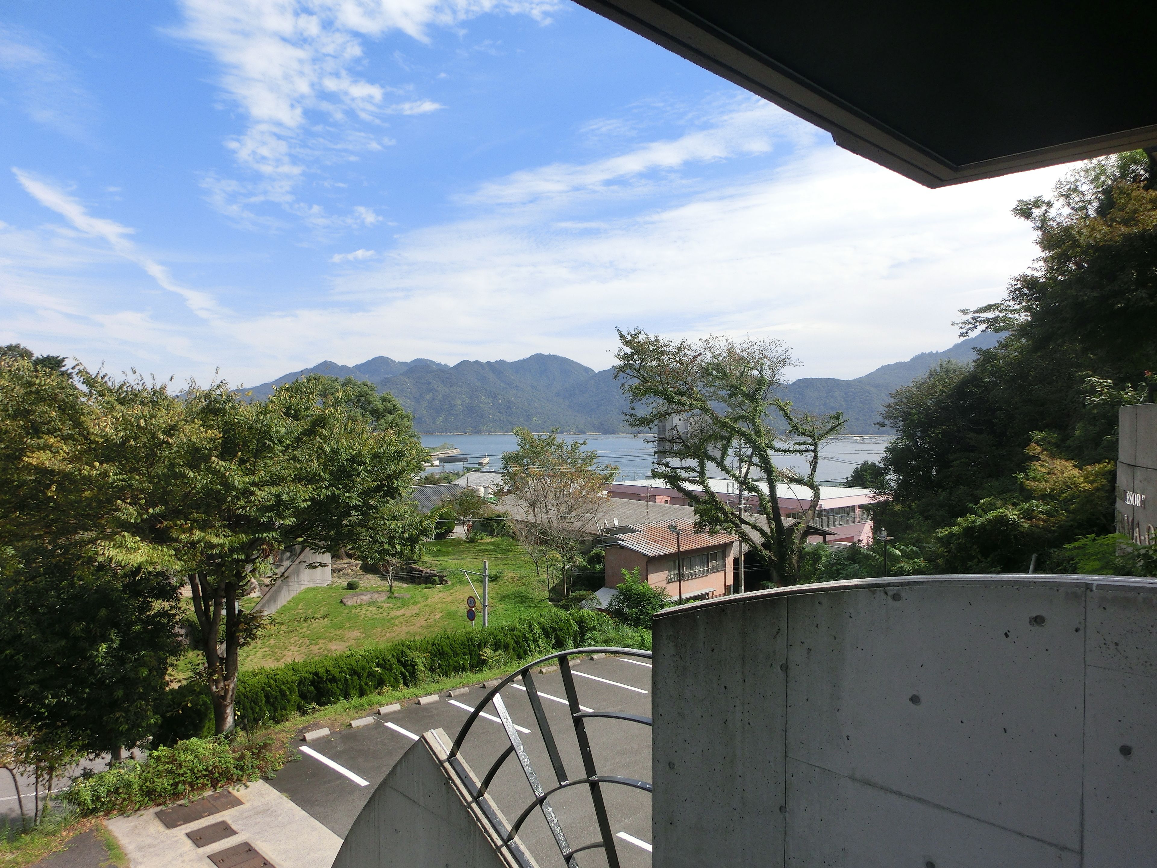 A view from a balcony overlooking a body of water, mountains, and a parking area with a building in the background.