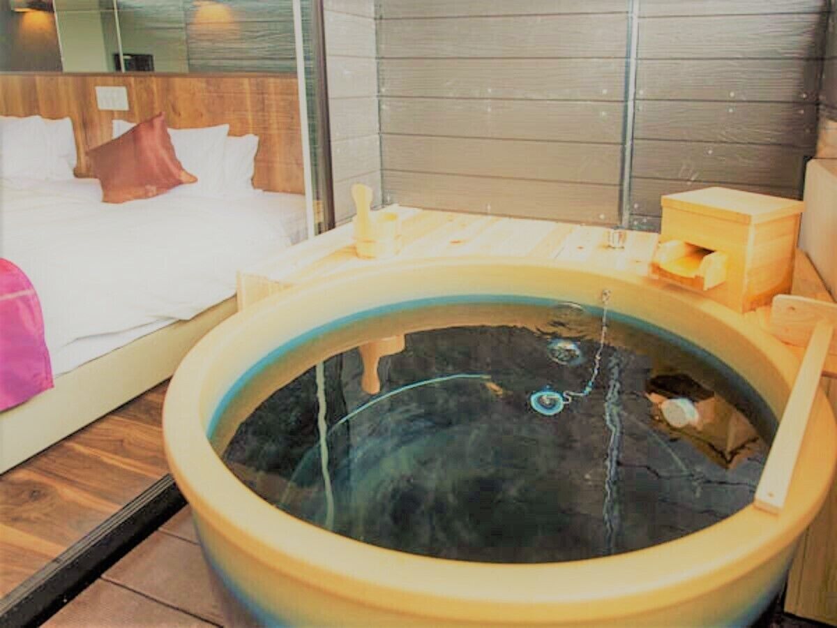 A room with a large bathtub, wooden headboard, and a bed with white linens and a pink pillow.